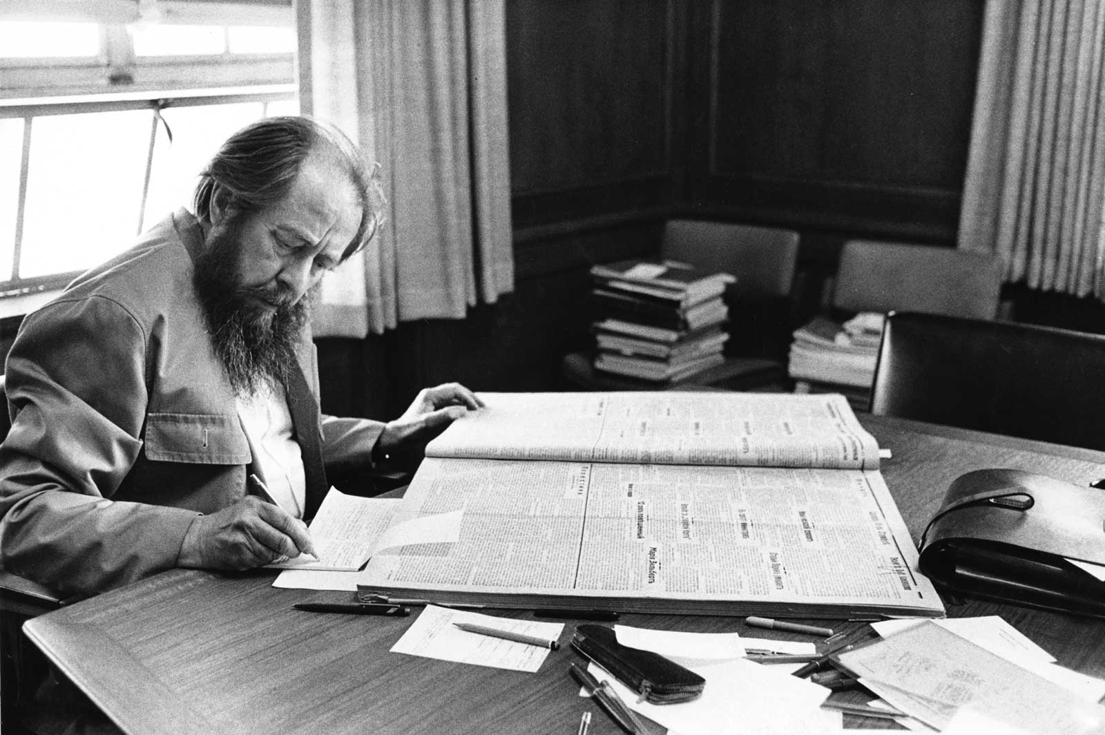 Aleksandr Solzhenitsyn sitting at a desk looking at newspapers