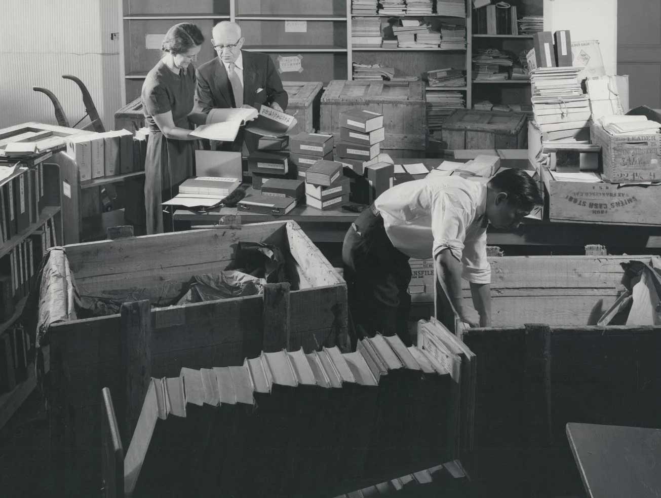 Black and white photo of 2 people standing looking at a book and 1 person searching through a box