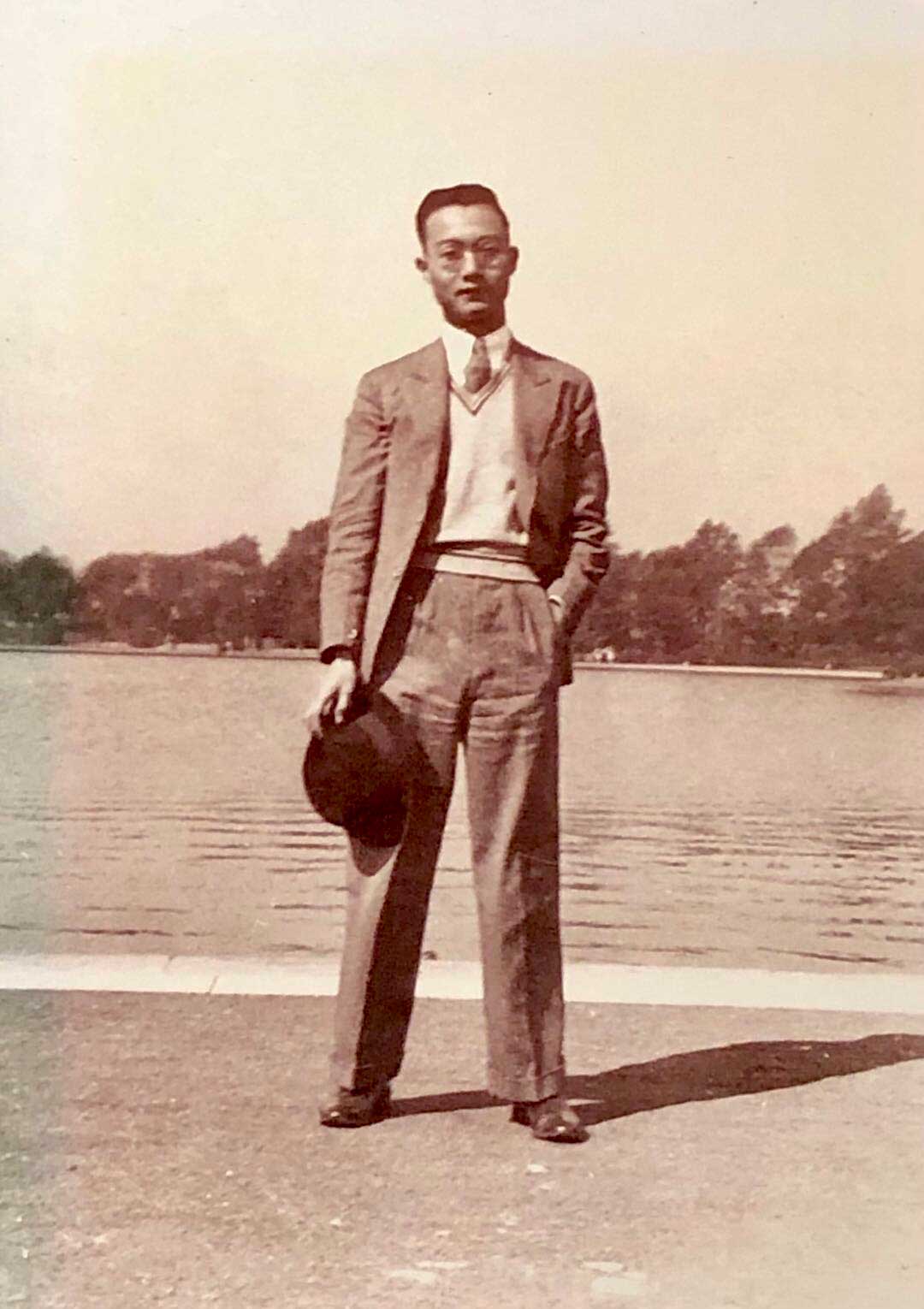 Chang You-ling wearing a suit holding a hat and standing in front of water with trees in the background
