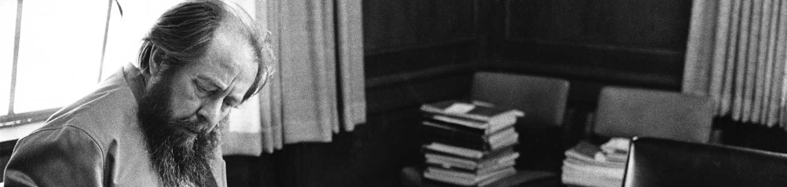 Aleksandr Solzhenitsyn sitting at a desk looking at newspapers