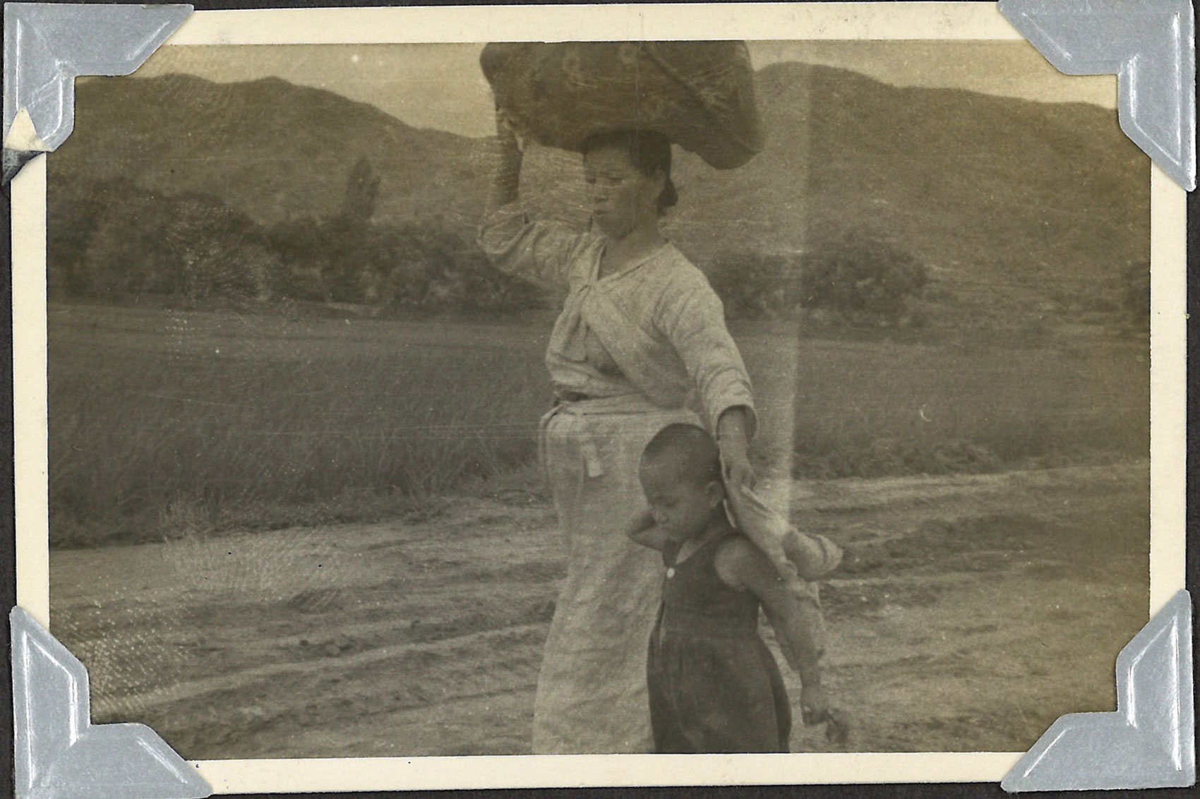 Black and white photo of woman carrying a bundle on top of her head next to a child.