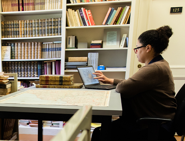 A researcher sits at right, working on a laptop with a large map unforlded at the table next to her and bookshelves in the background.