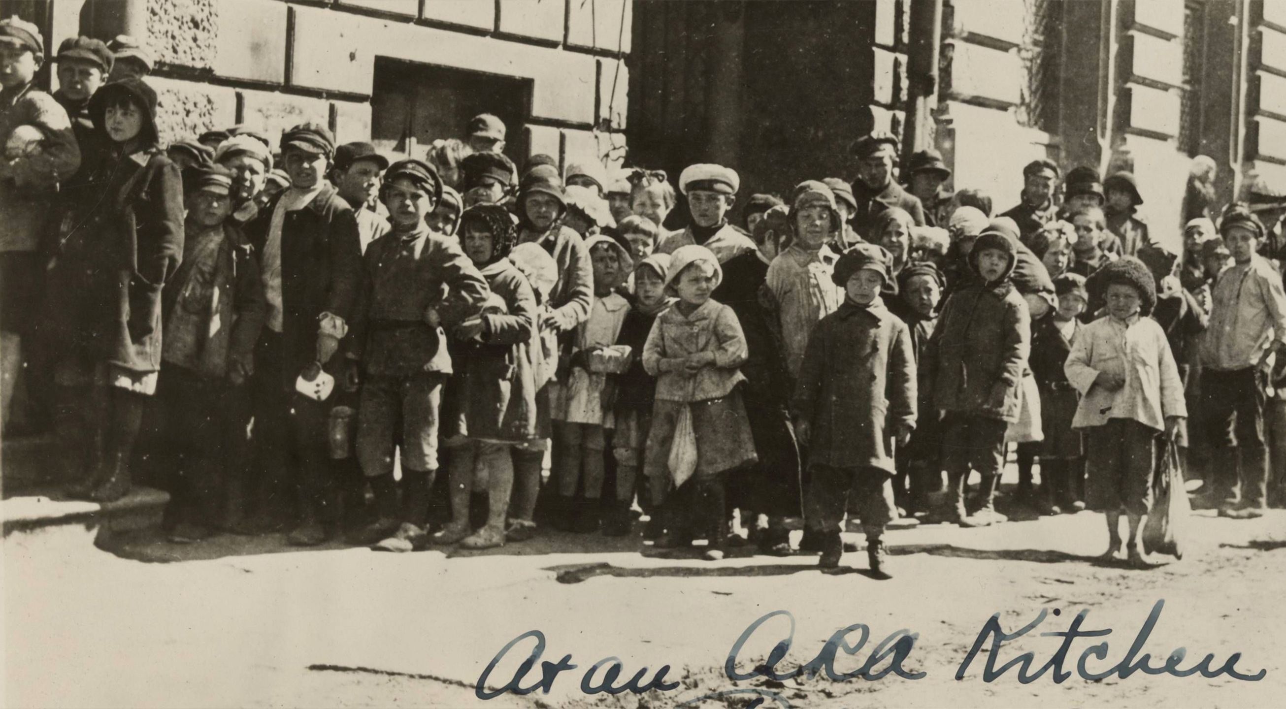 Black and white photograph showing a long line of children standing outside of an ARA kitchen in Petrograd, many clearly carrying their ration cards or knapsacks.