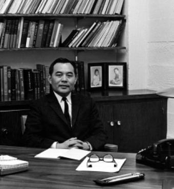 Black and white photo of Eugene Wu sitting at a desk with a bookshelf in the background