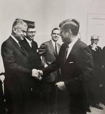 Black and white photo of 5 men in a room. Norman Obbard shaking hands with President John F. Kennedy.