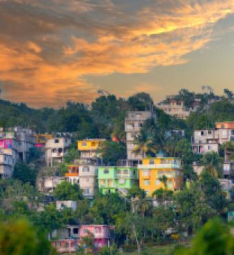 View of colorful houses on hilly area of Jamaica with lush foliage and a cloudy sky.