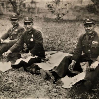 Black and white photo of 3 men dressed in military uniforms sitting on the ground having a picnic.