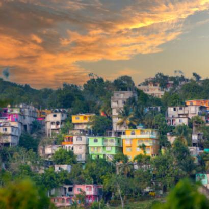 View of colorful houses on hilly area of Jamaica with lush foliage and a cloudy sky.