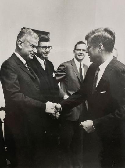 Black and white photo of 5 men in a room. Norman Obbard shaking hands with President John F. Kennedy.
