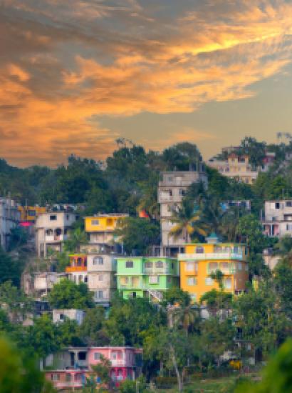 View of colorful houses on hilly area of Jamaica with lush foliage and a cloudy sky.