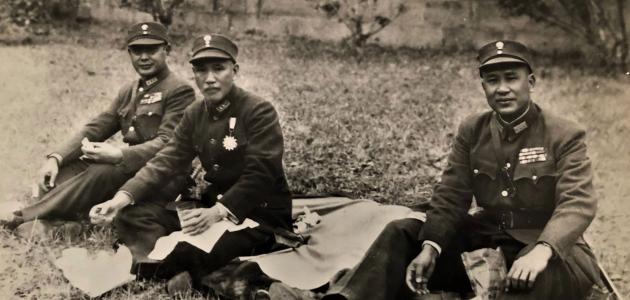 Black and white photo of 3 men dressed in military uniforms sitting on the ground having a picnic.