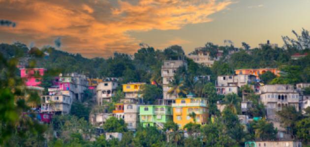 View of colorful houses on hilly area of Jamaica with lush foliage and a cloudy sky.