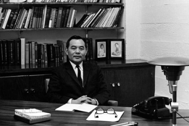 Black and white photo of Eugene Wu sitting at a desk with a bookshelf in the background