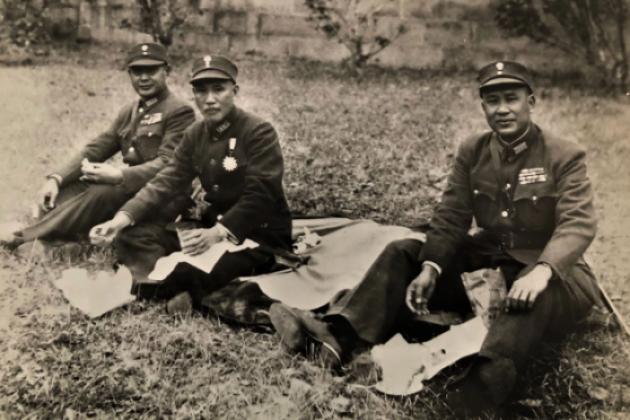 Black and white photo of 3 men dressed in military uniforms sitting on the ground having a picnic.