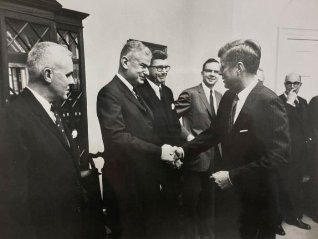 Black and white photo of 5 men in a room. Norman Obbard shaking hands with President John F. Kennedy.