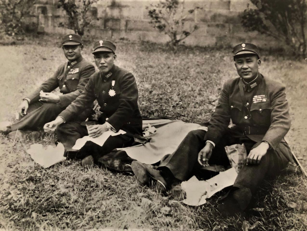 Black and white photo of 3 men dressed in military uniforms sitting on the ground having a picnic.