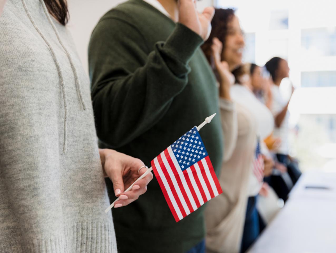 The group of multiracial people stand and raise their hand to say the pledge.