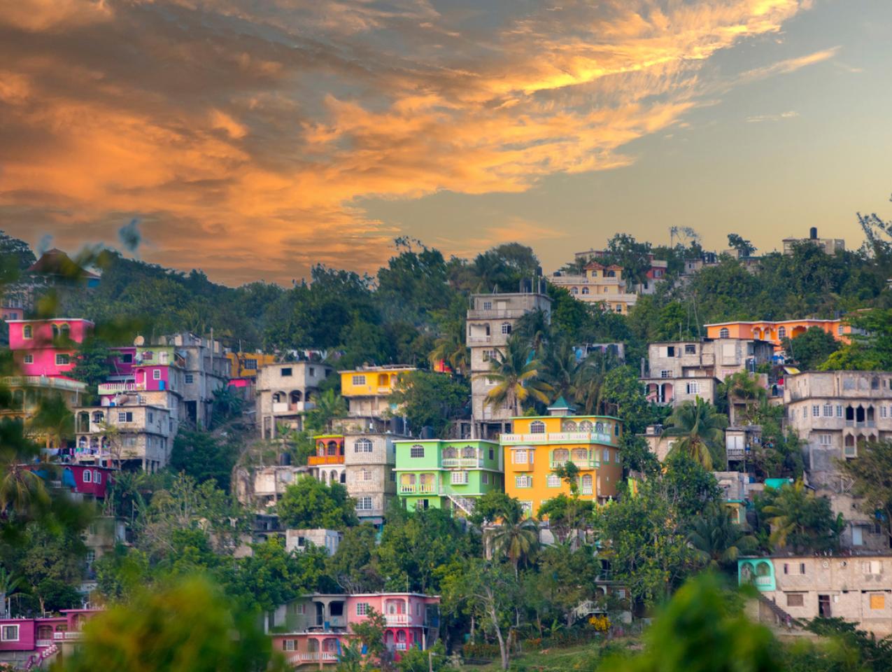 View of colorful houses on hilly area of Jamaica with lush foliage and a cloudy sky.