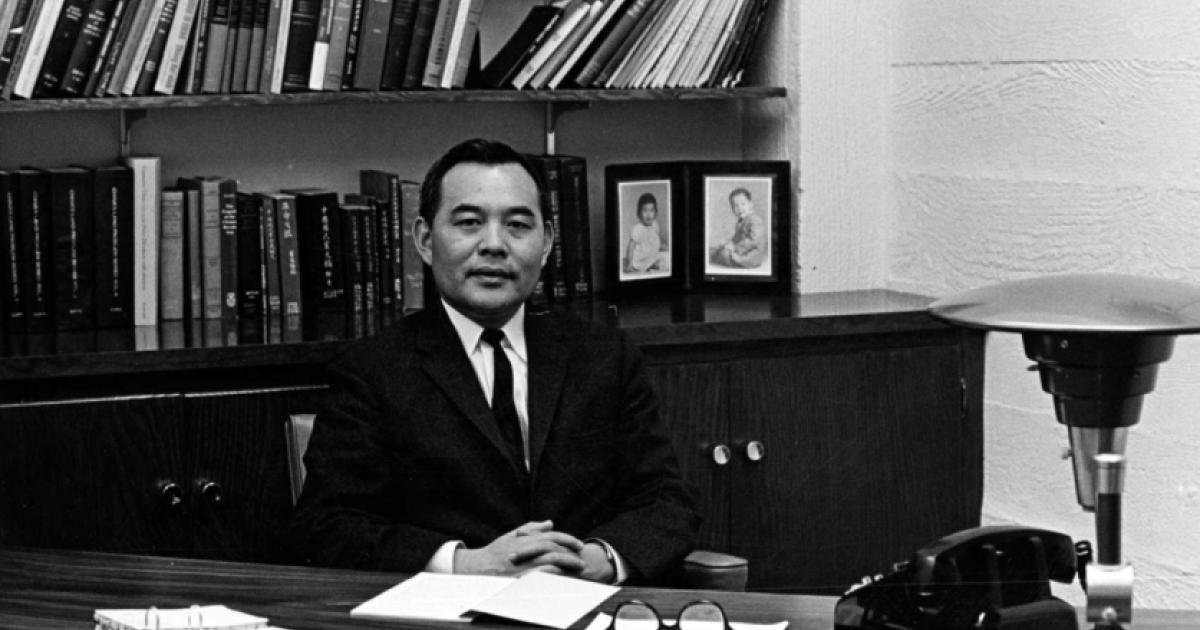 Black and white photo of Eugene Wu sitting at a desk with a bookshelf in the background