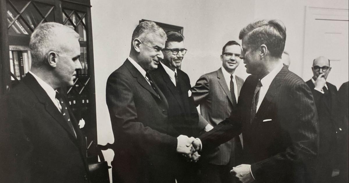 Black and white photo of 5 men in a room. Norman Obbard shaking hands with President John F. Kennedy.
