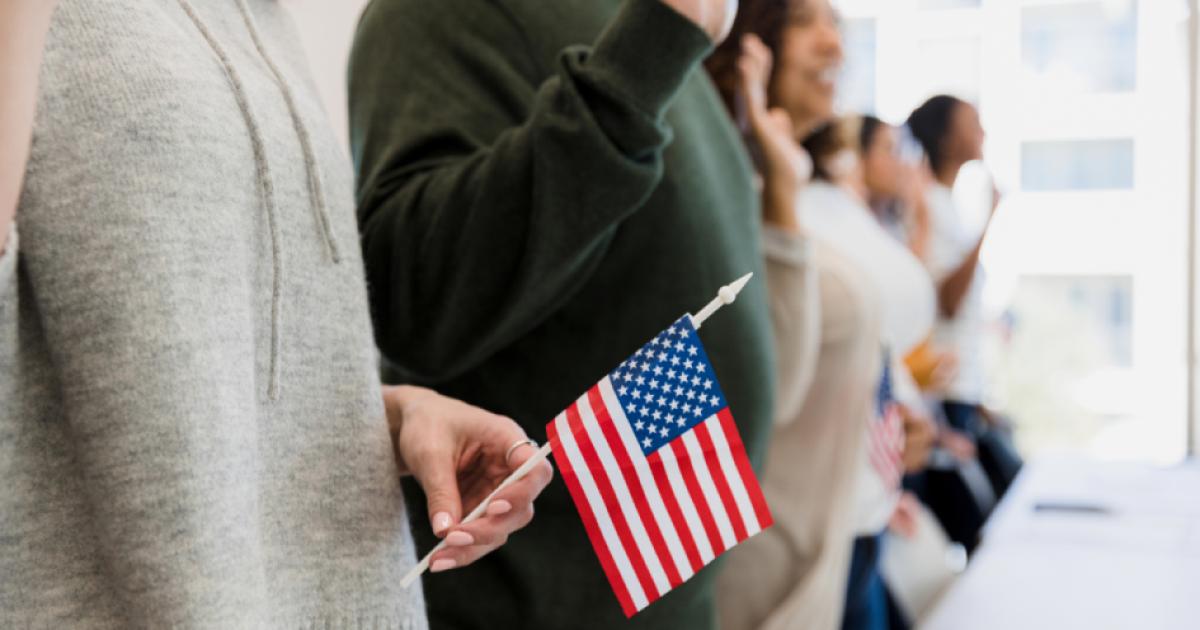 The group of multiracial people stand and raise their hand to say the pledge.