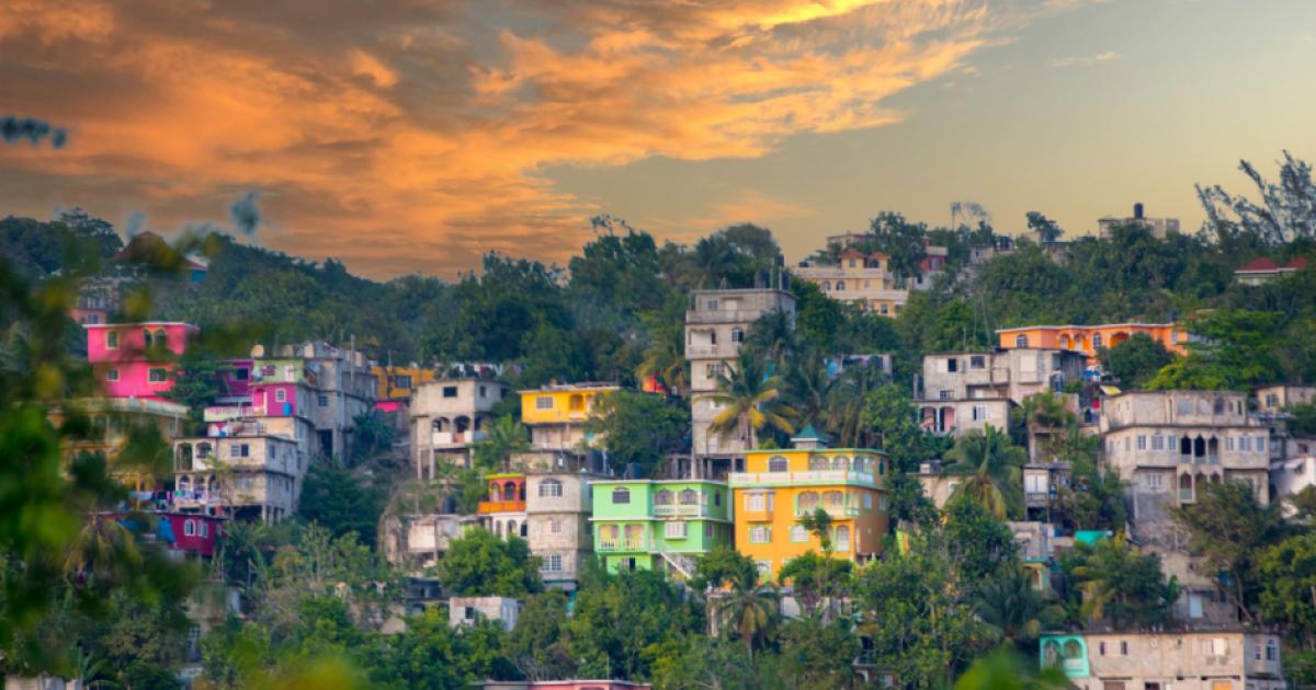 View of colorful houses on hilly area of Jamaica with lush foliage and a cloudy sky.